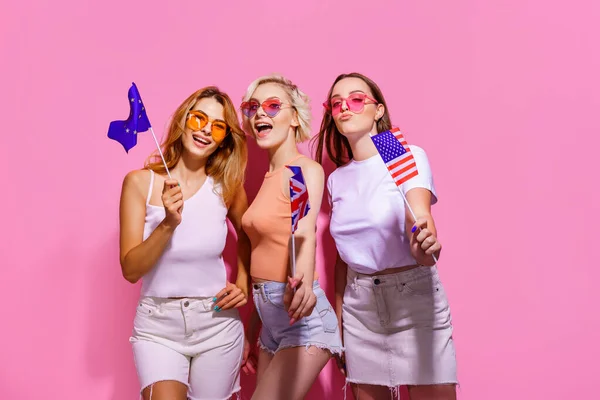 Tři dívky v letních šatech, brýle s vlajkami Evropské unie, Spojených států a Británie a pózují na růžovém pozadí ve studiu. — Stock fotografie