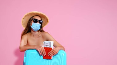 Uçuşunu bekleyen genç bir kadın, bavulunun yanında oturuyor, Coronavirus 'u önlemek için maske takıyor.