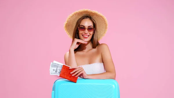 Pozitivní mladá dívka v slaměném klobouku a sluneční brýle s lístky a kufřík růžové studio pozadí, úsměvy při pohledu do kamery — Stock fotografie
