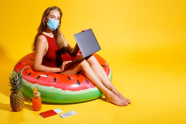 Jeune femme vêtue de maillot de bain rouge assise sur un cercle gonflable sur le fond jaune. portant un masque avec un ordinateur portable près des billets d'ananas cocktail regardant la caméra — Photo