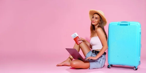 Glimlachend meisje met laptop tickets geld en paspoort gaan reizen. Zittend in de buurt van koffer in korte broek witte top en strohoed. Roze achtergrond. kijkt weg — Stockfoto