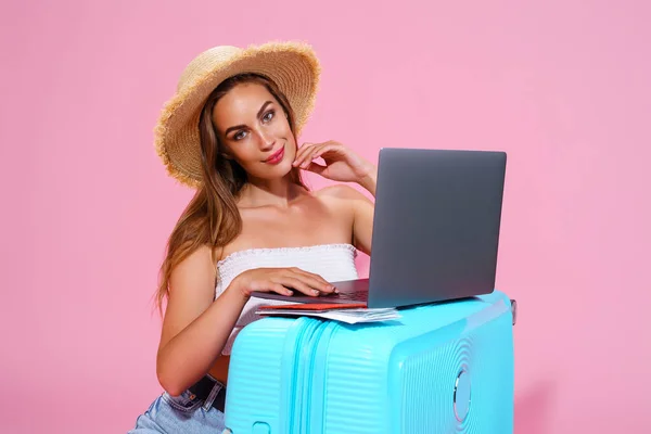 Glimlachend meisje met laptop tickets geld en paspoort gaan reizen. Portret. Zittend in de buurt van koffer in korte broek witte top en strohoed. Roze achtergrond. kijkt naar de camera — Stockfoto