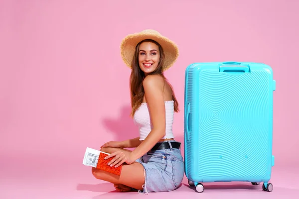 Улыбающаяся девушка с билетами собирается путешествовать. Сидя рядом с чемоданом в шортах и соломенной шляпе. Розовый фон — стоковое фото