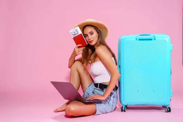 Glimlachend meisje met laptop tickets geld en paspoort gaan reizen. Zittend in de buurt van koffer in korte broek witte top en strohoed. Roze achtergrond. kijkt naar de camera — Stockfoto