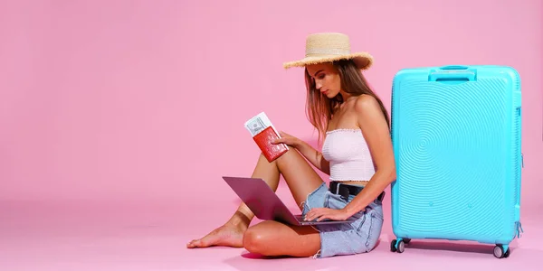 Meisje met laptop tickets geld en paspoort gaan reizen. Zittend in de buurt van koffer in korte broek witte top en strohoed. Roze achtergrond. Ik kijk naar de laptop. Iets zoeken. — Stockfoto