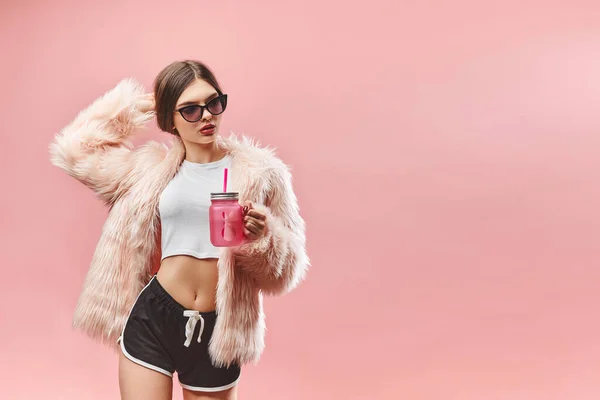 Девушка в искусственно розовом меховом пальто черные шорты белый топ и солнечные очки держит розовый напиток. С закрытыми глазами. Студия. Розовый фон. Красные губы. Брюнетка — стоковое фото