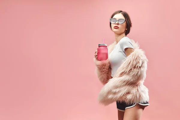 Meisje in faux pink bontjas zwarte korte broek witte top en zonnebril houdt roze drank. Met gesloten ogen. Een studio. Roze achtergrond. Rode lippen. Brunette — Stockfoto
