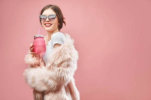 Chica sonriente en falso abrigo de piel rosa pantalones cortos negros top blanco y gafas de sol sostiene la bebida rosa. Con los ojos cerrados. Estudio. Fondo rosa. Los labios rojos. Morena. —  Fotos de Stock