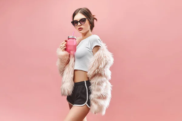 Chica en falso abrigo de piel rosa pantalones cortos negros top blanco y gafas de sol sostiene la bebida rosa. Con los ojos cerrados. Estudio. Fondo rosa. Los labios rojos. Morena. — Foto de Stock