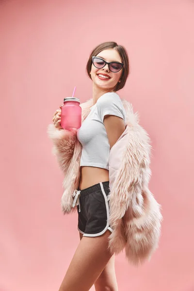 Chica sonriente en falso abrigo de piel rosa pantalones cortos negros top blanco y gafas de sol sostiene la bebida rosa. Con los ojos cerrados. Estudio. Fondo rosa. Los labios rojos. Morena. — Foto de Stock