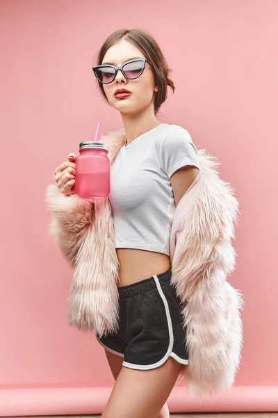 Chica en falso abrigo de piel rosa pantalones cortos negros top blanco y gafas de sol sostiene la bebida rosa. Con los ojos cerrados. Estudio. Fondo rosa. Los labios rojos. Morena. —  Fotos de Stock
