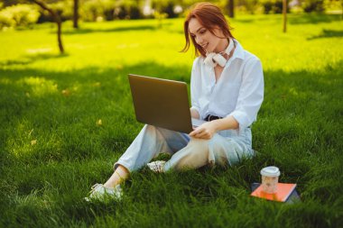 Parktaki güzel, genç, güzel, kızıl saçlı kadın çalışmak için dizüstü bilgisayar kullanıyor.