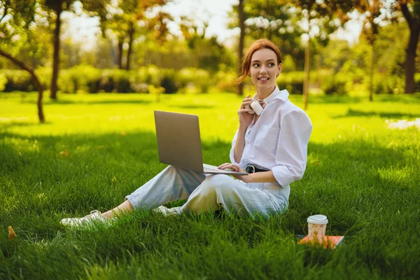 Красива молода красива руда жінка в парку на відкритому повітрі використовує ноутбук для навчання або роботи в Інтернеті, бездротові навушники на шиї — стокове фото