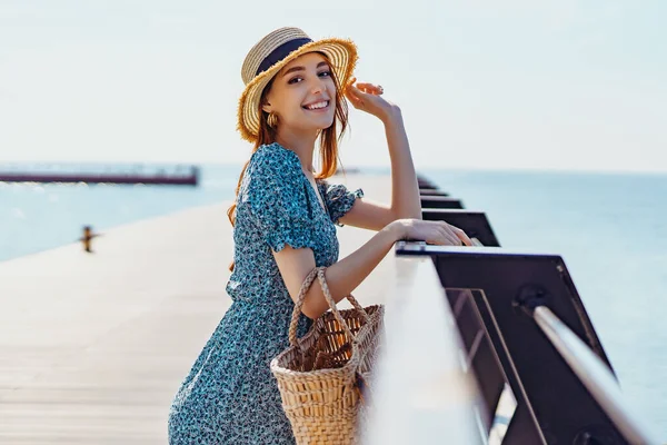Красива руда жінка позує в сонячний день. ходить уздовж пірсу біля моря. Дівчина в модній блакитній сукні та солом'яному капелюсі. Тримає солом'яну сумку. Дивлячись на камеру — стокове фото