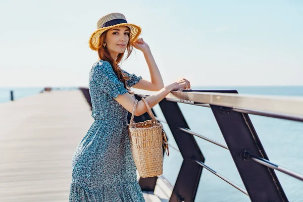 Mulher ruiva bonita posando no dia ensolarado. caminha ao longo do cais perto do mar. Menina vestindo óculos de sol na moda vestido azul e chapéu de palha. Segurando bolsa de palha — Fotografia de Stock