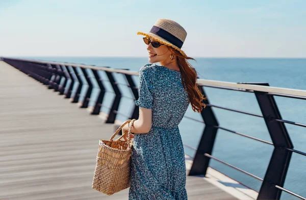 Красива руда жінка позує в сонячний день. ходить уздовж пірсу біля моря. Дівчина в модних сонцезахисних окулярах синя сукня і солом'яний капелюх. Тримає солом'яну сумку. Повертається і посміхається — стокове фото