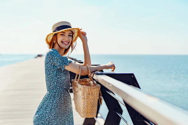 Красива руда жінка позує в сонячний день. ходить уздовж пірсу біля моря. Дівчина в модній блакитній сукні та солом'яному капелюсі. Тримає солом'яну сумку. Посміхаючись. Сміється. Дивлячись на камеру — стокове фото