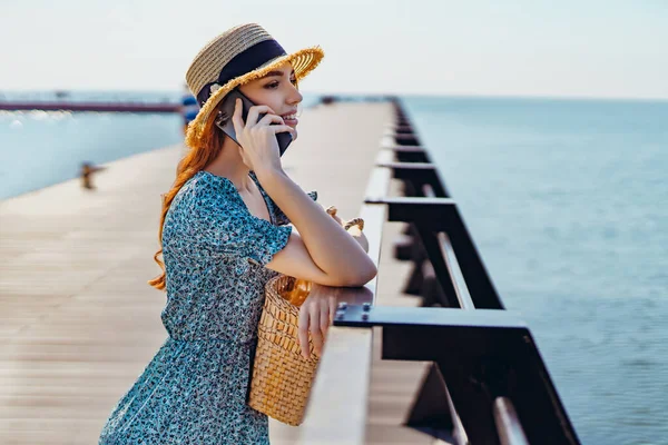 Ung smuk rødhåret pige taler på smartphone, mens du står på molen. Iført fashionable kjole halm hat og gyldne øreringe. Holding halm håndtaske og solbriller. Kigger væk - Stock-foto