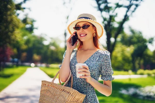 Alkalmi fiatal nő ruha szalma kalap és divatos napszemüveg beszél a telefonon, és kávét iszik, miközben séta kívül nyári zöld park kezében szalma táska és kávé menni. — Stock Fotó
