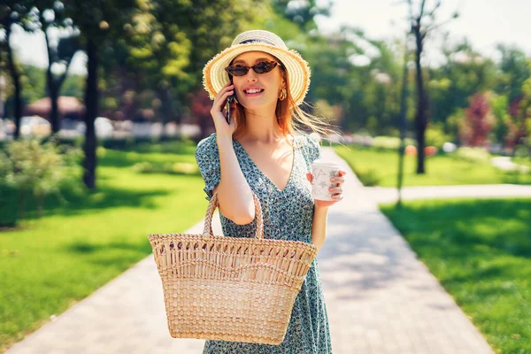 Jeune femme décontractée en robe chapeau de paille et lunettes de soleil à la mode parler au téléphone et boire du café tout en se promenant à l'extérieur dans le parc vert d'été tenant sac à main de paille et café à emporter. — Photo