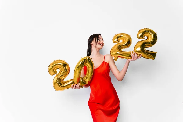 Joven mujer sonriente en vestido de cóctel rojo con maquillaje brillante celebrando Año Nuevo 2022 y sosteniendo globos dorados 2022 en manos sobre fondo blanco — Foto de Stock