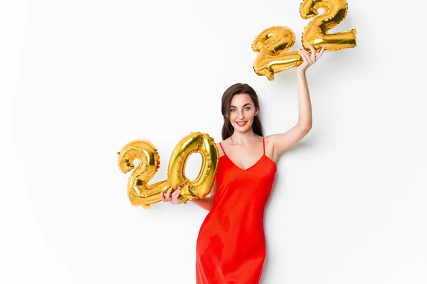 Jovem mulher sorridente em vestido de cocktail vermelho com maquiagem brilhante celebrando Ano Novo 2022 e segurando balões dourados 2022 em mãos no fundo branco — Fotografia de Stock