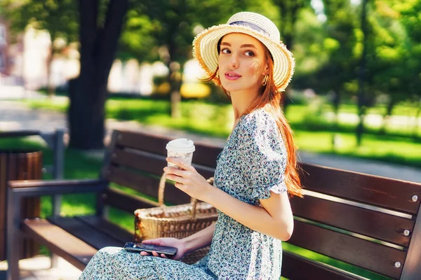 Gyönyörű vörös hajú fiatal nő pihen a padon a városi parkban, kezében mobiltelefon és kávé a kezében. Ruha szalmakalap viselése divatos fülbevalók — Stock Fotó
