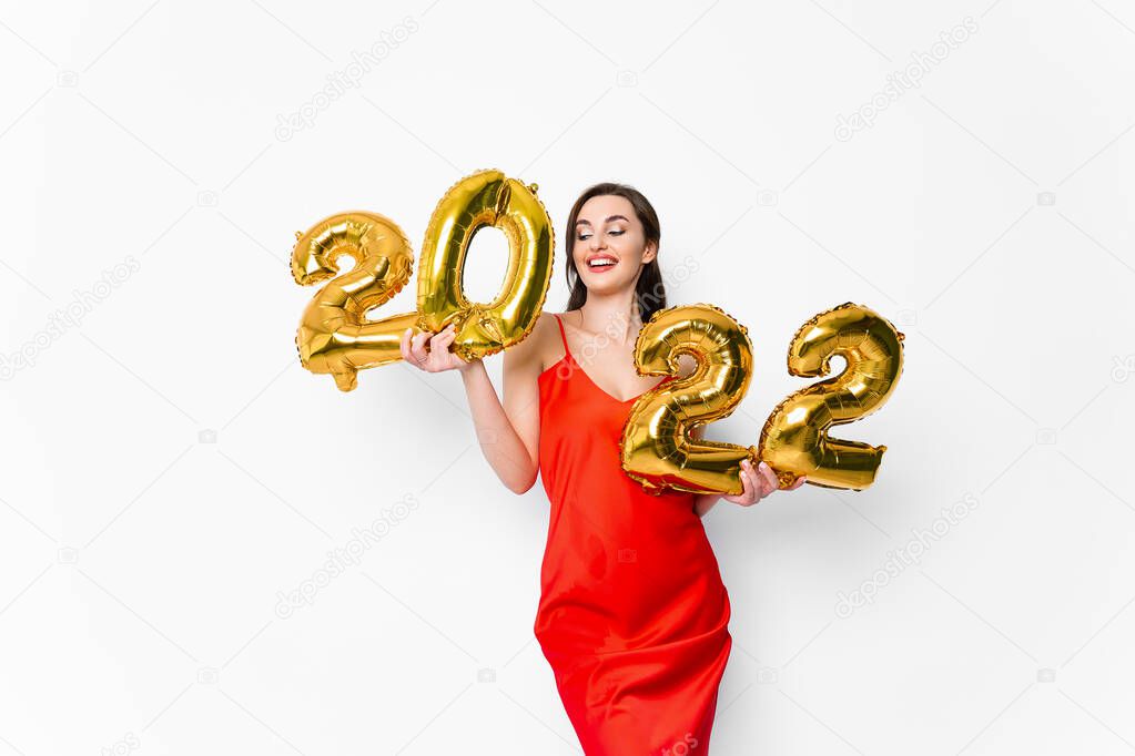 Young smiling woman in red cocktail dress with bright make-up celebrating New Year 2022 and holding golden balloons 2022 in hands on white background