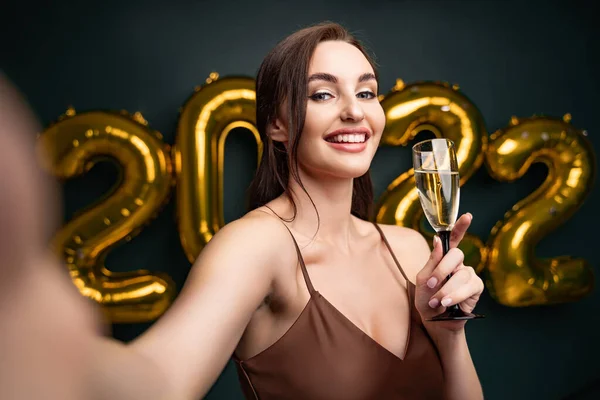 Schöne Frau im modernen Luxuskleid macht Selfie mit Champagner vor goldenen Luftballons vor dunklem Hintergrund — Stockfoto