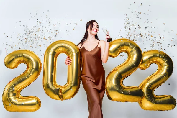 Jovem morena feliz com copo de champanhe perto de ouro 2022 balões no fundo branco com olhos fechados. Celebração do Ano Novo Confetti. Vestido de glamour na moda — Fotografia de Stock
