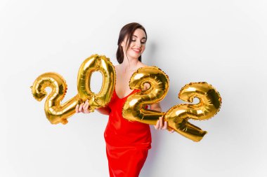 2022 yılını kutlayan parlak makyajlı, kırmızı elbiseli, gülümseyen genç bir kadın ve elinde 2022 altın balonları beyaz arka planda ellerinde tutuyor.