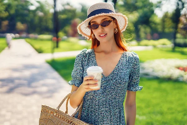 Молода красива дівчина посміхається, гуляючи в парку, тримаючи солом'яну сумку і каву в одноразовій чашці. Носіння в модних сонцезахисних окулярах синя сукня і солом'яний капелюх — стокове фото