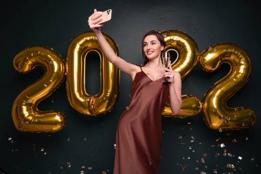 Lüks, modern elbiseli güzel bir kadın altın 2022 balonlarının önünde şampanyayla selfie çekiyor. Arka planda izole edilmiş.