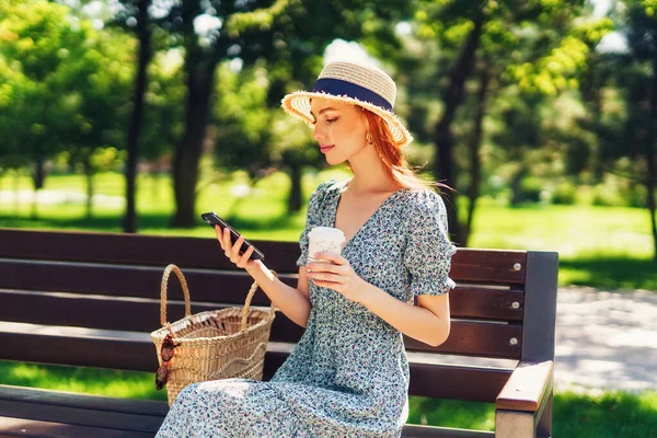 Красива руда молода жінка, що розслабляється на лавці в міському парку, тримає каву в руках, використовуючи телефон. Носіння в сукні солом'яний капелюх модні сережки — стокове фото