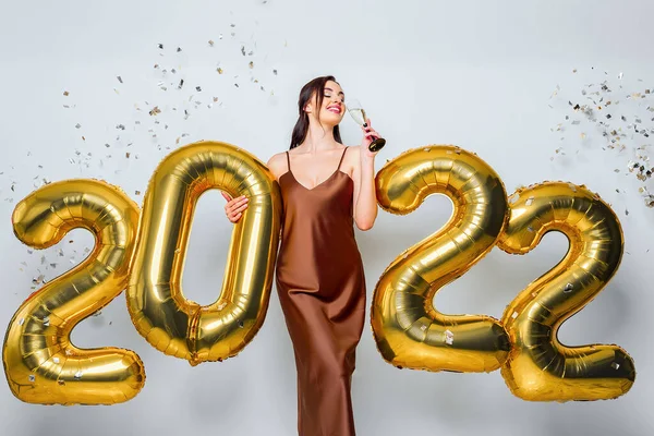 Jovem morena feliz com copo de champanhe perto de ouro 2022 balões no fundo branco. Celebração de Ano Novo. Vestindo vestido de glamour na moda. Confetti background. Beber champanhe — Fotografia de Stock