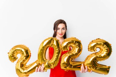 2022 yılını kutlayan parlak makyajlı, kırmızı elbiseli, gülümseyen genç bir kadın ve elinde 2022 altın balonları beyaz arka planda ellerinde tutuyor.
