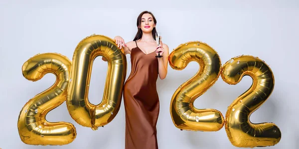 Jovem morena feliz com copo de champanhe perto de ouro 2022 balões no fundo branco. Celebração de Ano Novo. Vestindo vestido de glamour na moda. Olhando para a câmera — Fotografia de Stock