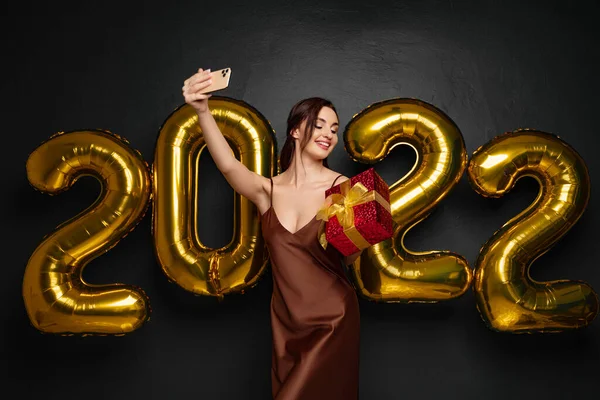 Sorrindo jovem morena em vestido elegante fazendo selfie no smartphone em fundo preto números dourados balões de ar. Olhando para a caixa de presente vermelha na mão. Feliz Ano Novo 2022 celebração feriado — Fotografia de Stock