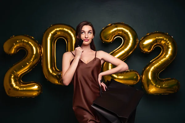 Woman in elegant dress hold credit bank card and shopping bags on black background, numbers air balloons studio.
