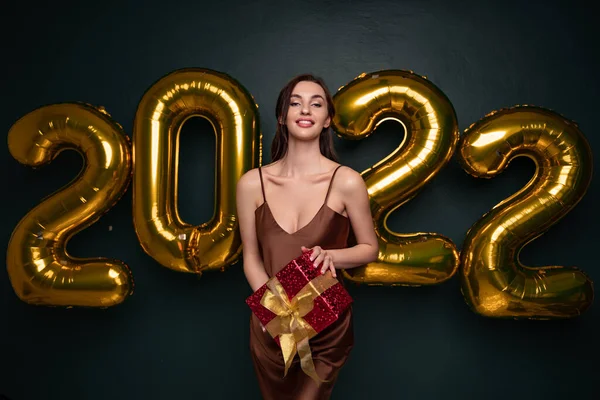 Жаркая брюнетка в красивом платье, держащая подарочную коробку на черном студийном фоне с золотыми воздушными шарами 2022 года, панорама. Счастливая молодая женщина с завернутым подарком на Новый год — стоковое фото