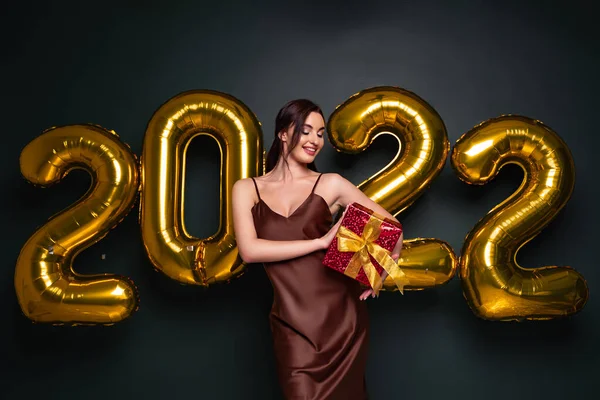 Жаркая брюнетка в красивом платье, держащая подарочную коробку на черном студийном фоне с золотыми воздушными шарами 2022 года, панорама. Счастливая молодая женщина с завернутым подарком на Новый год — стоковое фото