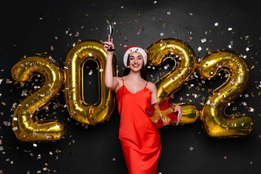 Yeni yıl partisi. Noel Baba şapkalı ve kırmızı elbiseli mutlu bir kadın şampanya kadehini kaldırıyor. Altın 2022 balon ve konfetiyle bayramı kutluyoruz. Kırmızı hediye kutusunu tutuyorum. Siyah arkaplan 