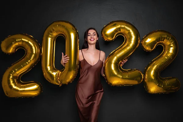 Jovem morena feliz em vestido glamour perto de ouro 2022 balões em fundo preto. Celebração de Ano Novo. Sorrindo. A olhar para a câmara. Rindo. Segurando números balões de ar — Fotografia de Stock