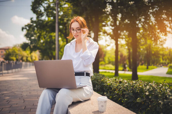 Beautiful redhead young woman sitting in the park and using laptop. Student. University. Freelance. Wearing in glasses jeans and white shirt. Coffee. Looking in laptop. adjusts glasses with hand 