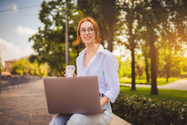 Beautiful redhead young woman sitting in the park and using laptop. Student. University. Freelance. Wearing in glasses jeans and white shirt. Holding in hand coffee to go. Looking away. Smiling