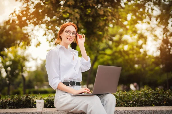 Красива руда молода жінка сидить у парку і використовує ноутбук. Студент. Університет. Фріланс. Носіння в окулярах повсякденних джинсів і білої сорочки. Кава, щоб піти. Паперова чашка. Дивлячись геть. усміхнений — стокове фото