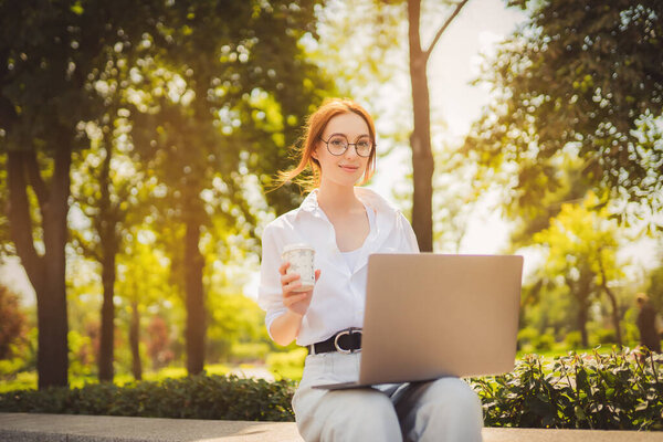 Beautiful redhead young woman sitting in the park and using laptop. Student. University. Freelance. Wearing in glasses jeans and white shirt. Holding in hand coffee. Looking at the camera. Smiling