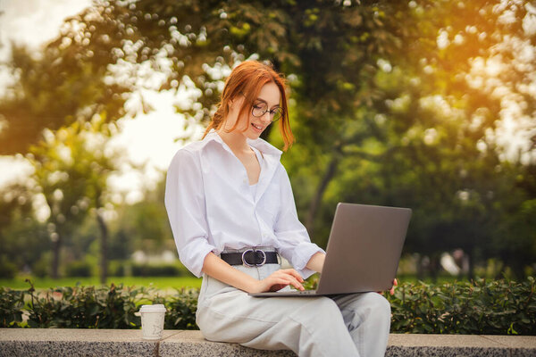 Beautiful redhead young woman sitting in the park and using laptop. Student. University. Freelance. Wearing in glasses casual jeans and white shirt. Coffee to go. Paper cup. Looking in laptop