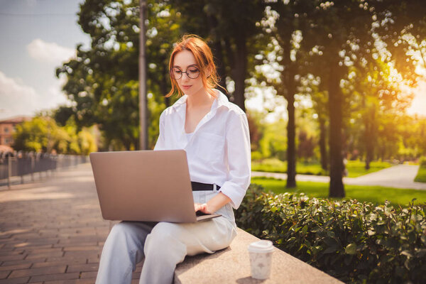 Beautiful redhead young woman sitting in the park and using laptop. Student. University. Freelance. Wearing in glasses casual jeans and white shirt. Coffee to go. Paper cup. Looking in laptop