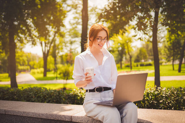 Beautiful redhead young woman sitting in the park and using laptop. Student. University. Freelance. Wearing in glasses jeans and white shirt. Holding in hand coffee. Looking at laptop. Smiling